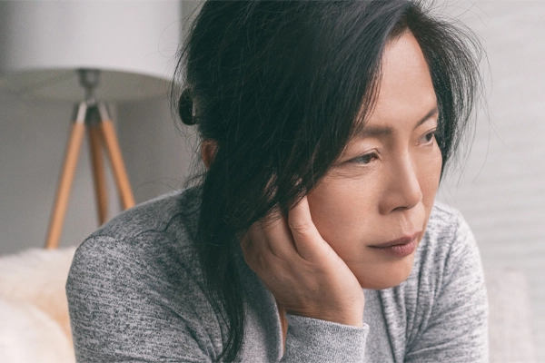 A middle-aged asian woman sitting on her couch, resting her head on her hand, suffering from adrenal fatigue. Get treatment for adrenal fatigue syndrome from Tanya Zboril, NP of TBT Medical in Ottawa–Gatineau.