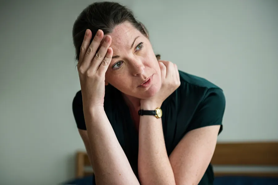 A woman in a dark blue dress sits on her bed holding her head in her hands with an exhausted expression. Get help for chronic fatigue syndrome from Tanya Zboril, NP of TBT Medical in [AERA].