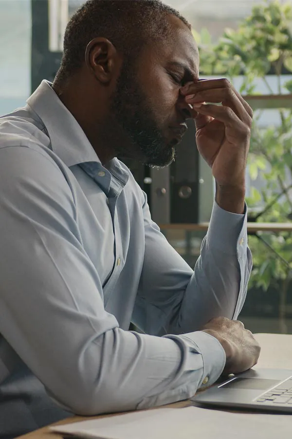 A black man rubbing the top of his nose at his desk, in need of chronic fatigue treatment from Tanya Zboril, NP of TBT Medical in Ottawa–Gatineau.