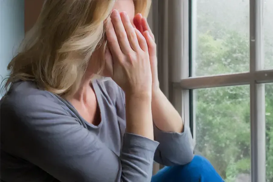 A blonde woman in a gray shirt sits in front of a window on a rainy day covering her eyes with her hands. Get treatment for Grave's Disease from Tanya Zboril, NP of TBT Medical in Ottawa–Gatineau.