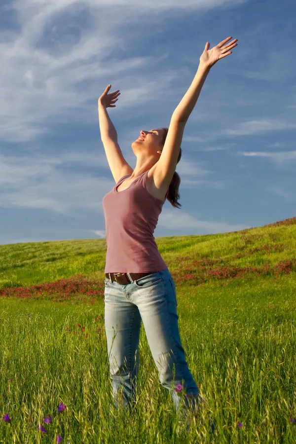 A woman in a tank top and jeans looks up with her arms outstretched to the sky in a field after successful treatment for Grave's Disease from Tanya Zboril, NP of TBT Medical in Ottawa–Gatineau.