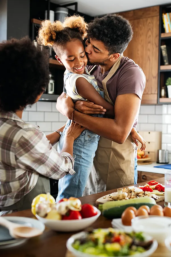 Gut-Health-Treatment A man holds his daughter and kisses her cheek as the family makes a healthy dinner in the kitchen. Get help for digestive issues and gut health from Tanya Zboril, NP of TBT Medical in Ottawa–Gatineau.