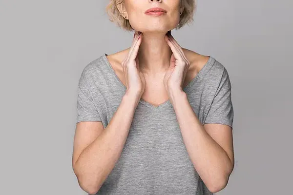 An older blonde woman in a gray t-shirt holds her hands to her neck checking for signs of thyroid problems. Get help for Hashimoto's Thyroiditis from Tanya Zboril, NP of TBT Medical in Ottawa–Gatineau.