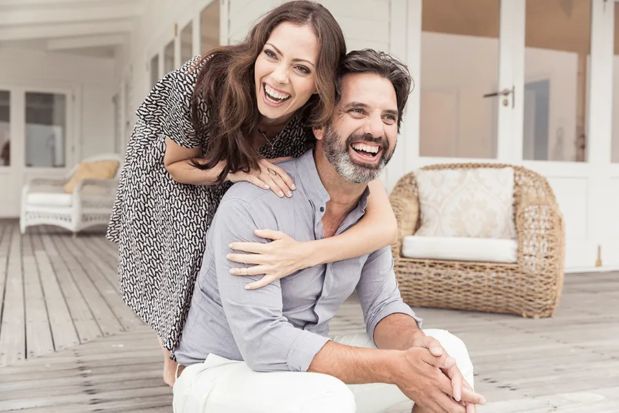 Health-and-Wellness-Services-Doctor A middle-aged couple on a wooden porch with wicker furniture; the woman is playfully hugging the man from behind. They are benefiting from the health and wellness services from Tanya Zboril, NP of TBT Medical in Ottawa–Gatineau.