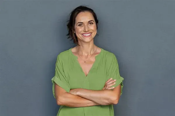 Hormones-for-Women-Doctor A woman in a green shirt stands smiling against a gray-blue wall, pleased with her hormone therapy from Tanya Zboril, NP of TBT Medical in Ottawa–Gatineau.