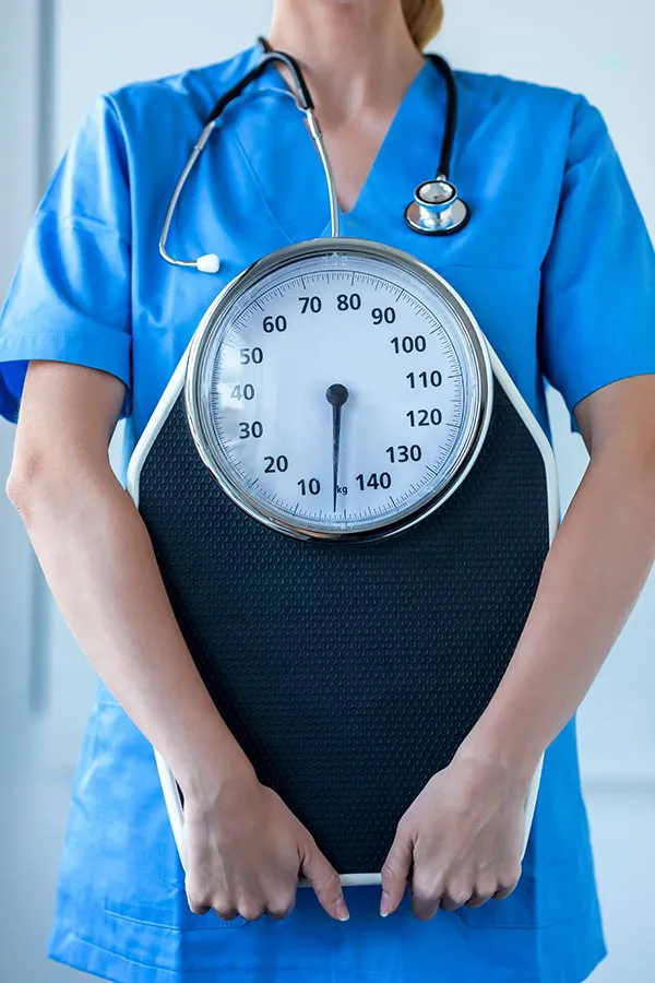 Medical-Weight-Loss-Treatment A nurse in blue scrubs with a stethoscope hanging around her neck holds a scale, representing weight loss treatments from Tanya Zboril, NP of TBT Medical in Ottawa–Gatineau.