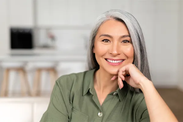 Menopause-Clinic An asian woman with long gray hair and a green shirt sitting and smiling on her couch, happy with her menopause care from [NAME} in Ottawa–Gatineau.
