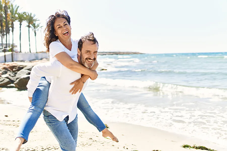 NAD-Therapy-Clinic A woman piggybacks on her husband as the middle-aged couple have fun at the beach. Get NAD Therapy from Tanya Zboril, NP of TBT Medical in Ottawa–Gatineau.