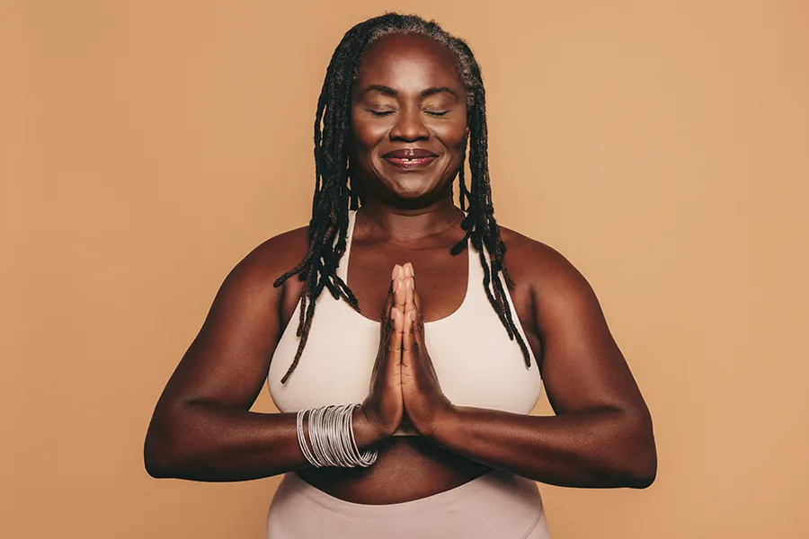 An older African American woman confidently posing against a warm background smiling with hands in a prayer pose. Tanya Zboril, NP of TBT Medical treats obesity using functional medicine in Ottawa–Gatineau.