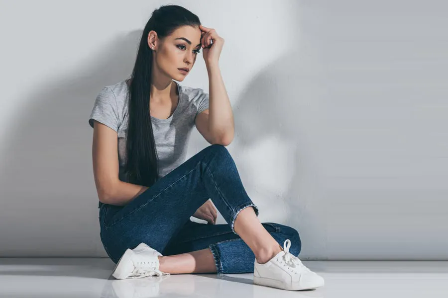A young brunette woman in a gray t-shirt and jeans sits against a gray wall with her head resting on her hand and a distressed expression. Get help for Polycystic Ovarian Syndrome (PCOS) from Tanya Zboril, NP of TBT Medical in Ottawa–Gatineau.