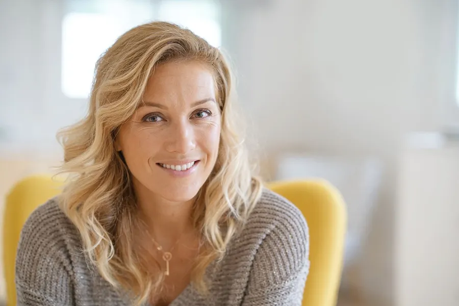 A blonde woman wearing a a gray sweater sits inside in a yellow chair, smiling and happy with her perimenopause treatment from Tanya Zboril, NP of TBT Medical in Ottawa–Gatineau.